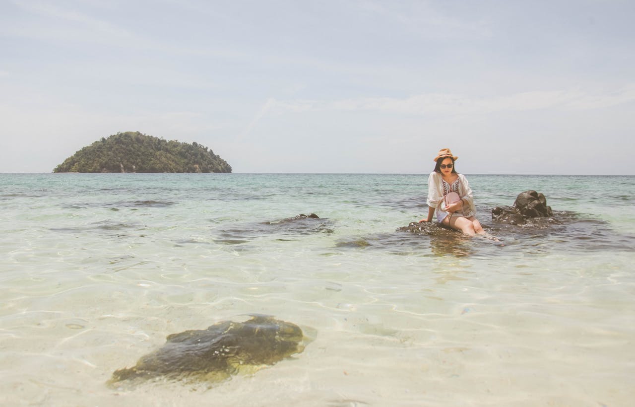 hero-img-01 Woman sitting on rocks by the sea with a hat, enjoying a tropical beach view.