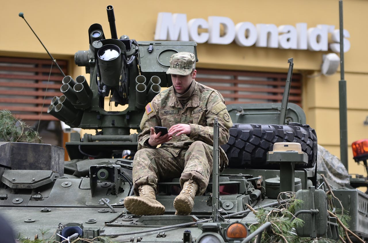 about-01 A soldier in camouflage resting on an armored vehicle, using a smartphone outside a McDonald's.